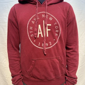 Vinröd hoodie från A&F med tryck - Snygg vinröd hoodie från Abercrombie & Fitch med stort rundat tryck på bröstet. Hoodien har huva med dragsko och en klassisk magficka. Perfekt för en avslappnad och trendig stil.