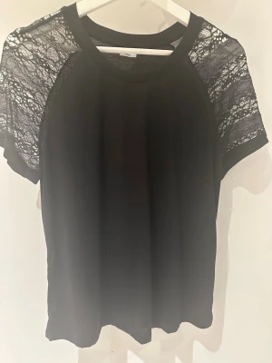 Svart topp med spets från Jacqueline de Yong storlek M - Snygg svart topp från Jacqueline de Yong med korta ärmar i transparent spets. Klassisk rund halsringning och mjukt material i kroppen. Perfekt för dig som vill ha en enkel men ändå detaljrik topp.