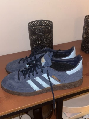 Blå adidas Spezial sneakers - Säljer ett par adidas Spezial sneakers i blå mocka med vita ränder och bruna gummisulor. Skorna har klassisk låg profil, snörning och guldfärgad Spezial-text på sidan. Perfekta för dig som gillar retrostil och streetwear.
