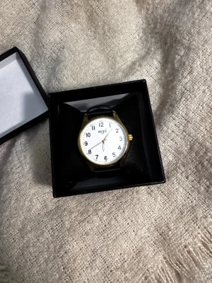 Regal klocka med svart läderband - Stilren klocka från Regal med vit urtavla, svarta siffror och guldfärgad boett. Klockan har ett klassiskt svart läderband och tydliga visare. Perfekt accessoar för en clean look. Helt oanvänd 