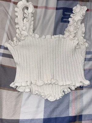 Vit stickad linnetopp med volang - Supersöt vit stickad linnetopp med volangdetaljer längs axelband och nederkant. Toppen har ribbad struktur och dekorativa pärlknappar framtill som ger en extra touch. Perfekt att styla till jeans eller kjol för en trendig look.