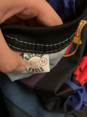 FC Barcelona Lamine Yamal tröja - Säljer en FC Barcelona matchtröja från Golden Eagle med Lamine Yamal och nummer 10 på ryggen. Tröjan har färgglada ränder i svart, rött och blått med gula detaljer och UNHCR-logga. Perfekt för dig som älskar fotboll och vill sticka ut på planen.