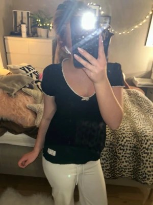 Svart topp med rosett från Odd Molly - Söt svart topp från Odd Molly med vit rosett och dekorativ vit kant runt v-ringningen. Kortärmad modell med lätt puffärm och mjukt bomullsmaterial. Liten etikett nertill på framsidan. .