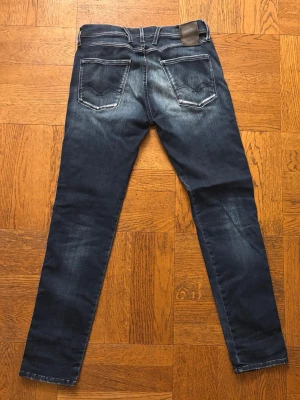Replay blå jeans med slitningar - Snygga blå jeans från Replay med slitningar och tvättade detaljer. Klassisk femficksmodell med raka ben och normal passform. Tydliga kontrastsömmar och Replay-logga i midjan. Perfekta för dig som gillar en avslappnad men stilren look.