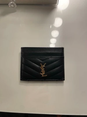 Svart YSL plånbok i skinn - Snygg svart plånbok från YSL i skinn med quiltad design och guldigt logotypemblem framtill. Perfekt för kort och sedlar, stilren och smidig att ha med sig. Klassisk och tidlös accessoar för dig som gillar lyxiga detaljer.