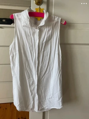 Vit ärmlös skjorta med krage - Klassisk vit skjorta utan ärmar med krage och knäppning framtill. Perfekt för en clean och enkel stil. Tillverkad i mjukt bomullstyg som känns skönt mot huden.