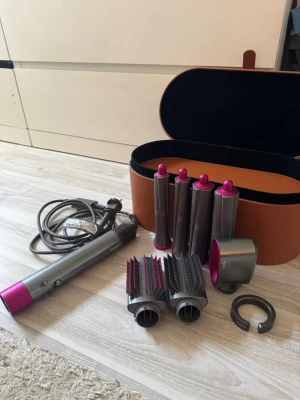 Dyson Airwrap hårstyler med tillbehör - Säljer en Dyson Airwrap hårstyler i silver och rosa med flera stylingtillbehör som borstar, locktänger och fönmunstycke. Allt kommer i ett snyggt brunt fodral. Perfekt för att skapa olika frisyrer med ett enda verktyg. Det går bra att använda köp nu💕