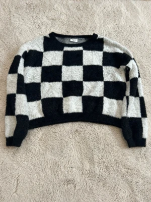 Svartvit rutig stickad tröja - Cool stickad tröja med svartvitt rutmönster och rund hals. Tröjan har en croppad passform och långa ärmar. Säljer pågrund av att jag växt ut den och knappt använt den