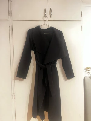 Svart lång kappa med bälte - Stilren svart kappa med bred krage och knytskärp i midjan. Lång modell med raka ärmar och klassisk siluett. Perfekt för dig som vill ha en enkel men snygg look till höst och vinter.