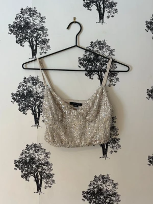 Glittrig croppad linnetopp - Snygg glittrig croppad linnetopp med tunna axelband och paljetter över hela toppen. Perfekt för dig som vill sticka ut och glänsa lite extra. Neutral beige färgton som passar till mycket och ger en lyxig känsla.