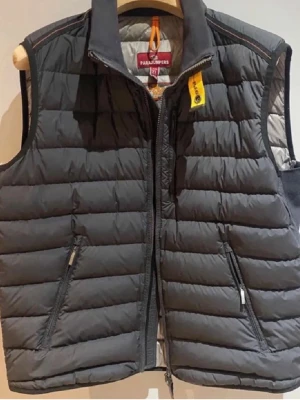 Svart dunväst från Parajumpers - Snygg svart dunväst från Parajumpers med quiltad design och dragkedja framtill. Västen har två snedställda fickor med dragkedja och en gul detalj vid kragen. Perfekt för lager-på-lager och håller dig varm under kyliga dagar.