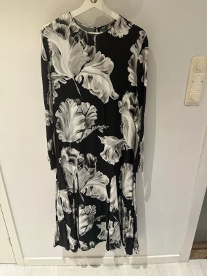 Svart långklänning med blommigt mönster storlek S - Svart långklänning med stora vita och grå blommor. Klänningen har lång ärm och rund halsringning. Tyget är mjukt och faller snyggt, perfekt för dig som gillar ett elegant och stilrent uttryck.