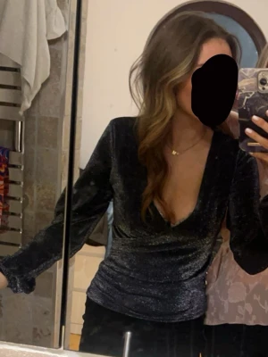 Glittrig omlottopp  - Supersnygg glittrig svart omlottopp med lång ärm och djup v-ringning. Toppen har en figurnära passform och är perfekt för att sticka ut. Materialet är mjukt och stretchigt, vilket gör den bekväm att bära hela kvällen. Endast använd en gång så som ny. 