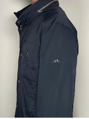 Bailey Poly Jacket J.Lindeberg - Snygg marinblå vindjacka från J.Lindeberg med fyra framfickor och diskret logga på ärmen. Jackan har hög krage, tryckknappar och dragkedja framtill. Tillverkad i slitstark polyester och passar perfekt till kyliga dagar.