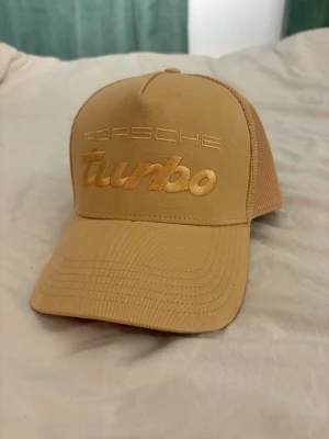 Beige Porsche Turbo keps - Snygg beige keps med broderad text 'Porsche Turbo' framtill. Kepsen har böjd skärm och mesh-paneler på sidorna och bak för extra ventilation. Perfekt accessoar för dig som gillar bilar och streetwear.
