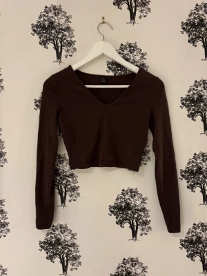 Brun croppad långärmad topp - Mörkbrun långärmad topp med croppad passform och v-ringning. Perfekt att styla med höga jeans eller kjol för en trendig look. Enkel och stilren design som passar till många outfits.