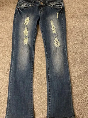 Jeans med slitningar - Snygga blå jeans med bootcut passform och slitna detaljer framtill. Jeansen har klassisk femficksdesign och knappgylf. Perfekta för en avslappnad och trendig look.