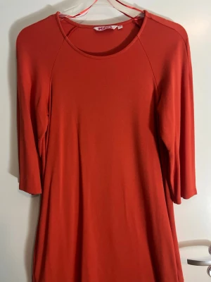 Röd/Orange klänning från Indiska - En enkel och stilren röd/orange klänning från Indiska med rund halsringning och trekvartsärm. Klänningen har en rak passform och är tillverkad i ett mjukt material som känns skönt mot huden. Perfekt för dig som gillar minimalistisk stil.