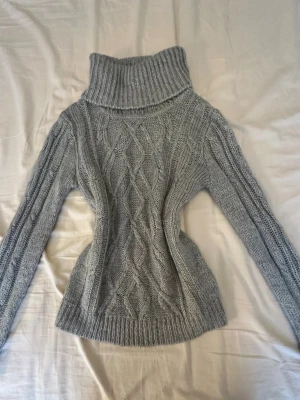Grå polotröja med kabelstickning - Snygg grå stickad polotröja med kabelstickat mönster framtill och ribbade muddar. Tröjan har lång ärm och hög, bred polokrage som ger en cool och mysig look. Perfekt för kalla dagar och enkel att matcha med jeans eller kjol.