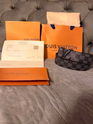 Louis Vuitton Damier bälte svart/grå - Säljer ett Louis Vuitton bälte i klassiskt Damier-mönster i svart och grått. Bältet har en stor LV-logga som spänne och är gjort i canvas med läderdetaljer. Perfekt accessoar för dig som gillar lyxiga detaljer och streetstyle. Kvitto allt ingår