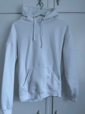 Vit hoodie med magficka - Klassisk vit hoodie med huva och dragsnören. Hoodien har en stor magficka framtill och ribbade muddar vid ärmslut och nederkant. Perfekt för en clean och avslappnad stil. Materialet känns mjukt och bekvämt, troligen bomullsblandning.