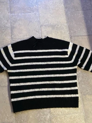 Svartvit randig stickad tröja H&M - Säljer en svartvit stickad tröja från H&M med breda horisontella ränder. Tröjan har en rak, boxig passform, ribbstickade muddar och rund hals. Perfekt för dig som gillar klassiska mönster och vill ha en mysig, trendig look.