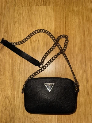 Svart axelväska från Guess med kedja - Snygg svart axelväska från Guess med silverfärgad kedjerem och klassisk trekantslogga framtill. Väskan har dragkedja upptill och ett ljust innerfoder med Guess-tryck samt kortfack. Perfekt storlek för det viktigaste och ger en trendig vibe.
