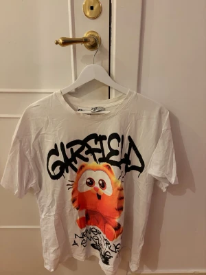 Vit Garfield t-shirt med tryck - Vit t-shirt med stort färgglatt Garfield-tryck och graffiti-inspirerad text på framsidan. 🚨det finns lite avfrågningar från smink runt halsen!! Se bild🚨