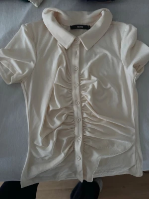 Beige kortärmad skjorta från Bik Bok - Snygg beige skjorta från Bik Bok med korta ärmar och klassisk krage. Skjortan har knappar framtill och cool rynkdetalj längs knappraden som ger en unik look. Tillverkad i mjukt material som känns skönt mot huden.