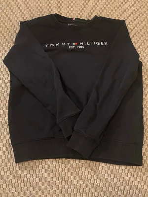 Svart sweatshirt från Tommy Hilfiger - Svart sweatshirt från Tommy Hilfiger med broderad logga och texten 'EST. 1985' på bröstet. Tröjan har rund halsringning och ribbade muddar vid ärmslut och nederkant. Perfekt för en avslappnad och stilren look. Storlek 164cm