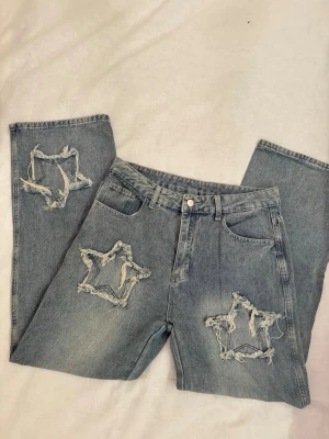 jeans med slitna stjärndetaljer - Unika grå jeans med stora slitna stjärnor på benen. Jeansen har klassisk femficksdesign, bälteshällor och knappgylf. Perfekta för dig som vill sticka ut med en cool och trendig look.
