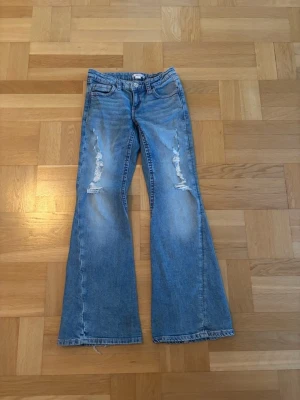 Blå bootcut jeans med broderade fickor - Säljer ett par ljusblå bootcut jeans med slitningar framtill och snygga broderade detaljer på bakfickorna. Jeansen har låg midja och är tillverkade i klassiskt denimtyg. Perfekta för dig som gillar en retro och avslappnad stil. 