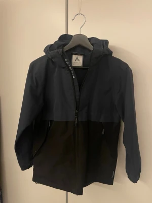 Regnjacka  - Stilren svart vindjacka från Mckinley med huva och hel dragkedja framtill. Jackan har två sidofickor med dragkedja och en enkel, clean look som funkar till allt. Tillverkad i lätt polyester och perfekt för regniga dagar.