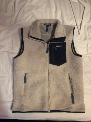 Patagonia Väst - Säljer en väst från Patagonia med marinblå detaljer och dragkedjor. Västen har hög krage, tre fickor med dragkedja och en kontrasterande bröstficka. Perfekt för lager-på-lager och riktigt mysig att bära.