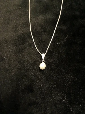 Silverhalsband med pärlhänge - Stilrent halsband i silver med ett elegant vitt pärlhänge. Kedjan är tunn och smidig, perfekt för att ge en klassisk touch till din look. Hänget har en droppformad infattning i silver som håller en rund, skimrande pärla.