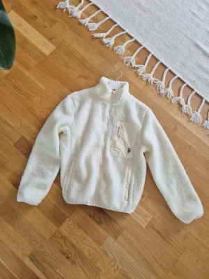 Vit piléjacka med dragkedja - Mysig vit teddyjacka med hög krage och dragkedja framtill. Jackan har en bröstficka med dragkedja och två sidofickor. Tillverkad i fluffigt teddy-material som håller dig varm och bekväm. Perfekt för kyliga dagar och enkel att matcha med olika outfits.