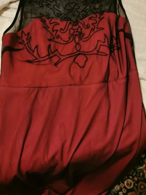 Vinröd klänning med spetsdetaljer - Säljer en vinröd klänning med transparent svart mesh upptill och broderade detaljer. Klänningen har en elegant design med vackra mönster på bröstet och är ärmlös. Perfekt för dig som vill sticka ut med en unik look.