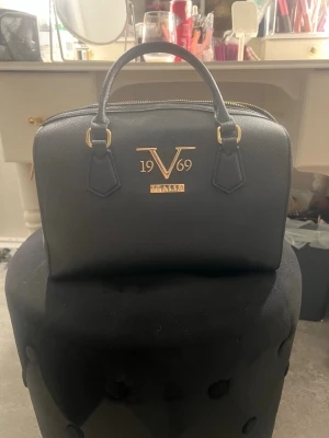 Svart handväska från V1969 Italia - Snygg svart handväska från V1969 Italia med guldfärgade detaljer och logga framtill. Väskan har två handtag och är tillverkad i ett strukturerat material som ger en exklusiv känsla. Perfekt för dig som vill ha en stilren och klassisk accessoar.