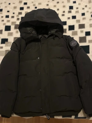 Svart pufferjacka från Canada Goose - Säljer en svart pufferjacka från Canada Goose med huva och patch på ärmen. Jackan har dragkedja och tryckknappar framtill samt praktiska innerfickor och bärremmar på insidan. Perfekt för kalla dagar och riktigt snygg streetstil. Kvitto finns. Priset kan diskuteras 