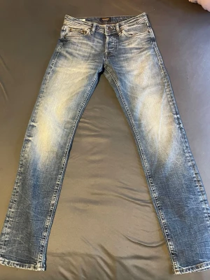Blå raka jeans från Jack & Jones - Säljer ett par blå jeans från Jack & Jones med klassisk femficksdesign och slitna detaljer framtill och baktill. Jeansen har normal passform och raka ben, tillverkade i jeansmaterial med snygg tvättad look och kontrastsömmar.