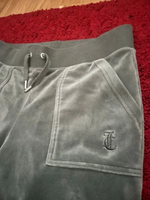 Juicy Couture Gröna Mjukisbyxor - Mysiga gröna mjukisbyxor i velour med bred resår i midjan och snörning. Byxorna har raka ben och två stora fickor framtill, varav ena är dekorerad med en broderad bokstav. Perfekta för chill dagar hemma.