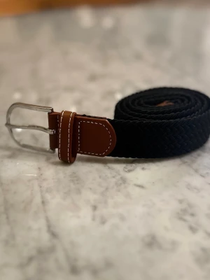 Flätat svart bälte med läderdetaljer - Snyggt flätat svart bälte med bruna läderdetaljer och vit kontrastsöm. Spännet är i silverfärgad metall och bältet har en flexibel, elastisk känsla. Perfekt accessoar för att lyfta din outfit.