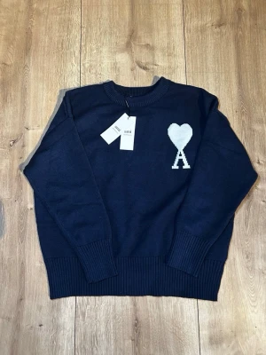 Mörkblå stickad tröja från Ami Paris - Mörkblå stickad tröja från Ami Paris med rund halsringning och ribbade muddar. På bröstet finns ett vitt hjärta med bokstaven A, vilket är Ami Paris ikoniska logga. Tröjan har en avslappnad passform och är perfekt för lager på lager.