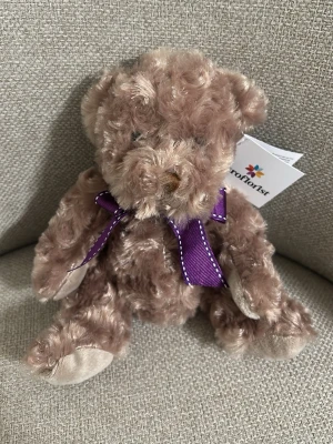 Mjuk brun nallebjörn med lila rosett - Söt och fluffig nallebjörn i ljusbrunt, lockigt plyschmaterial. Nallen har en lila rosett runt halsen och mjuka, ljusbeiga tassar. Perfekt som present eller dekoration. Märkt med Euroflorist-tag.