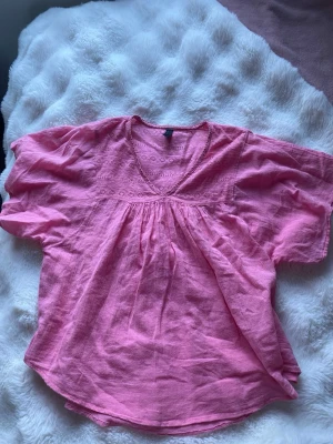 Rosa vid blus med broderi - Söt rosa blus med v-ringning och broderade detaljer upptill. Blusen har en lös och luftig passform med korta, vida ärmar. Perfekt för en avslappnad och trendig look. Materialet känns lätt och bekvämt mot huden.