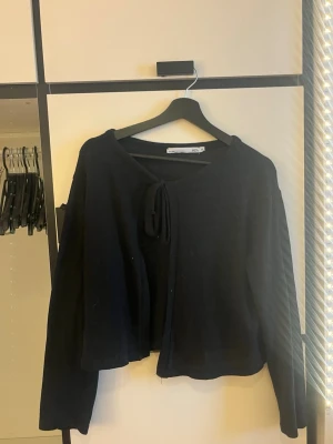 Svart stickad kofta med knytband - Snygg svart stickad kofta från 157 med knytband framtill. Koftan har lång ärm och en avslappnad, lite croppad passform. Perfekt att slänga över en topp för en chill look. Enkel och stilren design som passar till det mesta.