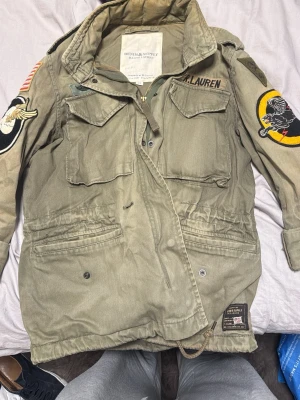 Grön parkajacka från Ralph Lauren - Säljer en militärinspirerad grön parkajacka från Ralph Lauren med flera coola patchar på ärmarna och bröstet. Jackan har dragkedja, knappar, fyra stora fickor framtill och justerbar midja. Perfekt för dig som gillar streetwear och utilitystil.
