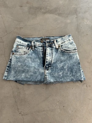 Blå jeanskjol från Colins - Säljer en kort jeanskjol från Colins i tvättad blå denim. Kjolen har rå kant nertill, fem fickor och bälteshällor. Klassisk knapp- och dragkedjestängning framtill. Perfekt för dig som gillar en avslappnad och trendig stil.