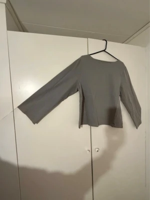 Grå långärmad topp med vida ärmar - En stilren grå topp med långa, vida ärmar och rund halsringning. Toppen har en rak passform och är tillverkad i ett mjukt bomullsliknande material. Perfekt att matcha med jeans eller kjol för en clean look.
