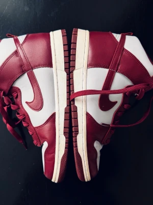 Nike Dunk High vinröd/ljusgrå sneakers - Nike Dunk High sneakers i vinrött och ljusgrått med klassisk hög siluett. Skorna har röd snörning, perforerad tåbox och ikoniska swoosh-loggan på sidan. Ovandelen är i skinn och syntet, vilket ger en stilren och sportig look.